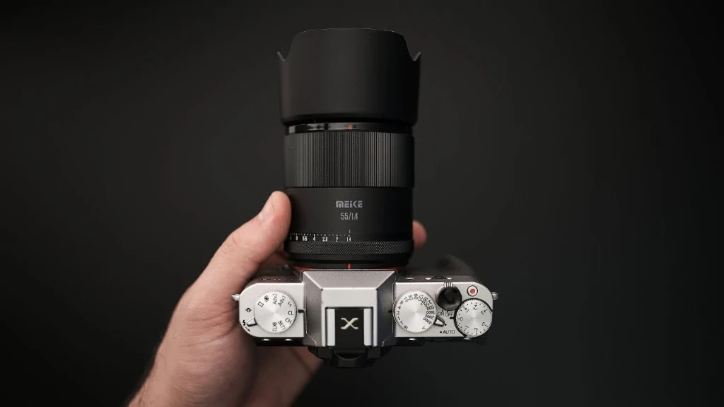 Светосильный объектив Meike AF 55mm f/1.4 с цилиндрической пластиковой блендой на камере Fujifilm в руке человека на черном фоне