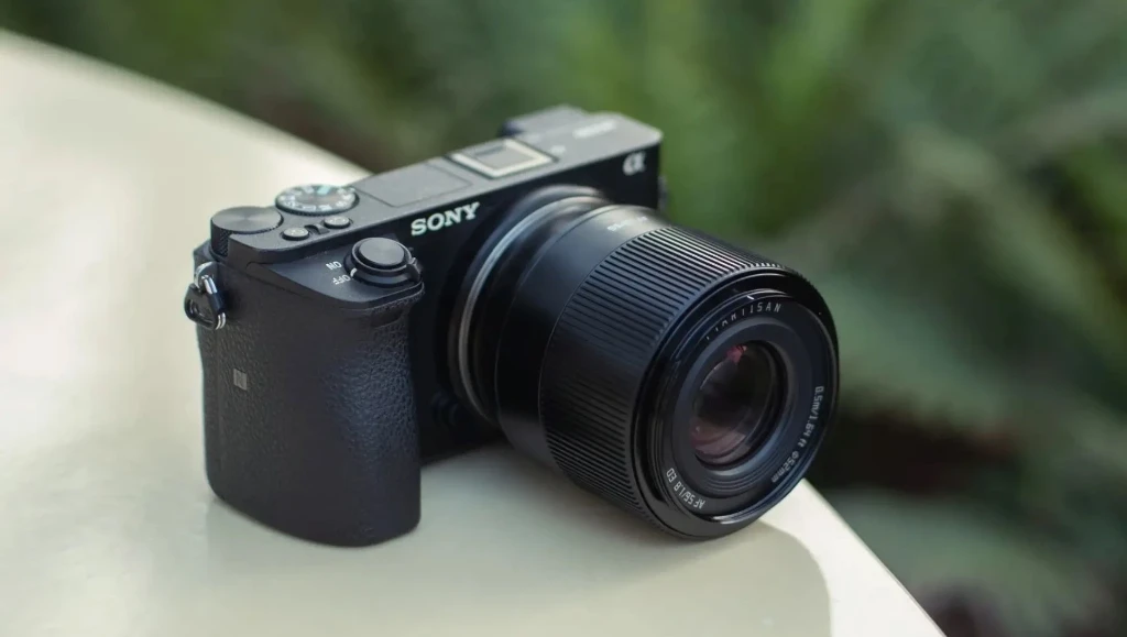Черный объектив TTArtisan AF 56mm f/1.8 в металлическом корпусе на беззеркальной камере Sony на наклонном парапете на фоне листвы