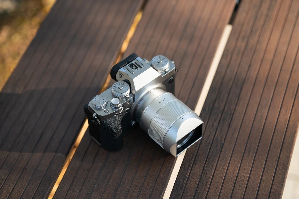 Серебристый металлический объектив TTArtisan AF 56mm f/1.8 с прямоугольной металлической блендой на серебристой камере Fujifilm на деревянном столе