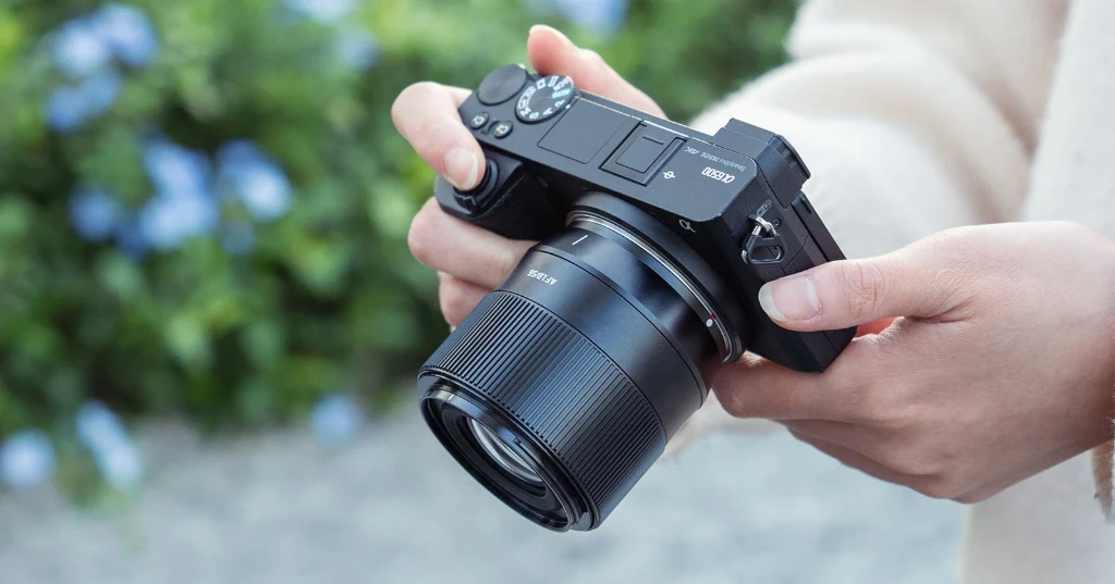 Черный объектив TTArtisan AF 56mm f/1.8 на камере Sony a6500 со встроенным стабилизатором SteadyShot в руках фотографа на улице