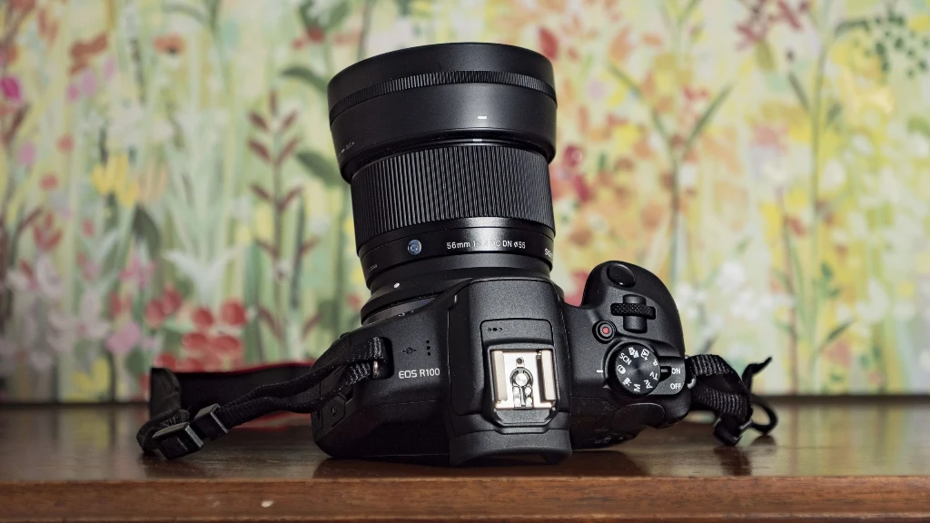 Черный объектив Sigma 56mm f/1.4 DC DN Contemporary с надетой цилиндрической блендой на компактной камере Canon EOS R100 в интерьере