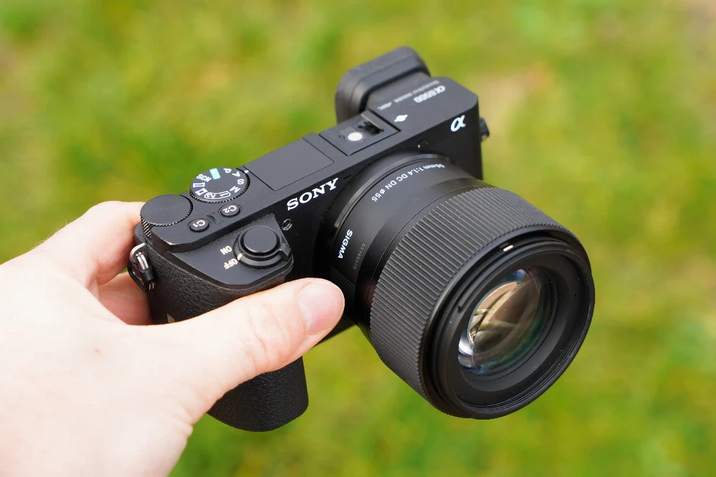 Объектив Sigma 56mm f/1.4 DC DN Contemporary без бленды на беззеркальной камере Sony a6500 с видоискателем в руке человека на фоне травы